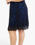 A close-up of a model wearing a knee-length black lace skirt with a vibrant blue floral lace overlay and scalloped black lace hem, set against a plain white background.