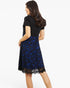 A woman shown from the back wearing a short-sleeve black top and a knee-length blue and black floral lace skirt against a plain white studio background.