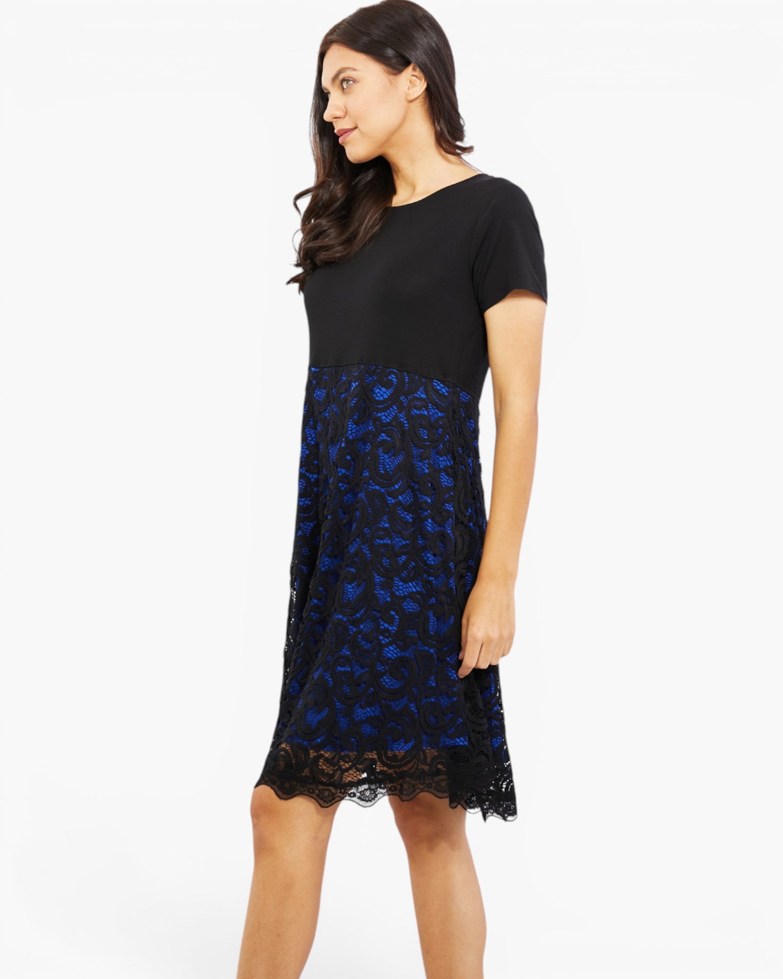 A woman modeling a short-sleeve knee-length dress with a solid black upper bodice and a black lace overlay skirt featuring a vibrant blue floral pattern, set against a plain white studio background.