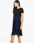 A woman modeling a short-sleeve knee-length dress with a solid black upper bodice and a black lace overlay skirt featuring a vibrant blue floral pattern, set against a plain white studio background.