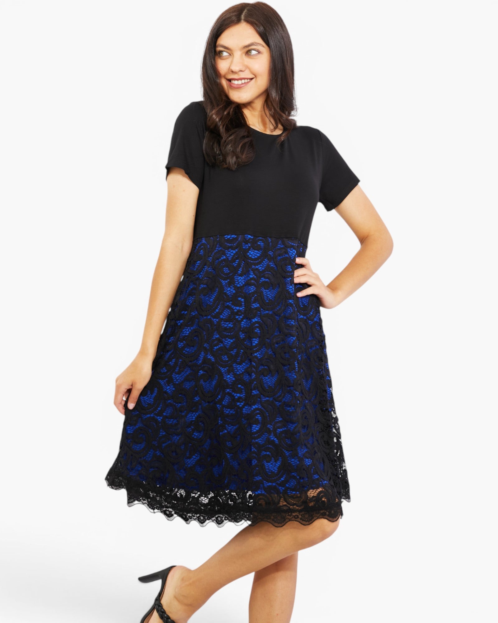 A woman modeling a short-sleeve midi dress with a solid black bodice and a full black lace skirt featuring a vibrant blue floral pattern, set against a plain white studio background.