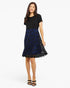 A woman modeling a short-sleeve knee-length dress with a solid black bodice and a black lace overlay skirt featuring a vibrant blue floral pattern, set against a plain white studio background.