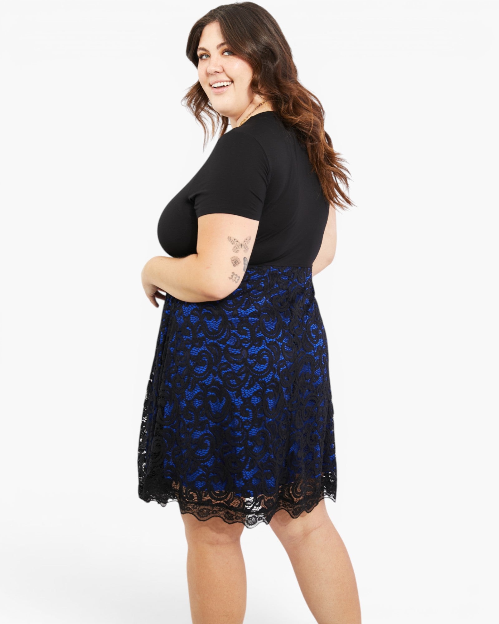 A woman modeling a black short-sleeve top and a black lace skater skirt with a royal blue floral pattern, shown from a three-quarter back angle against a white studio background.