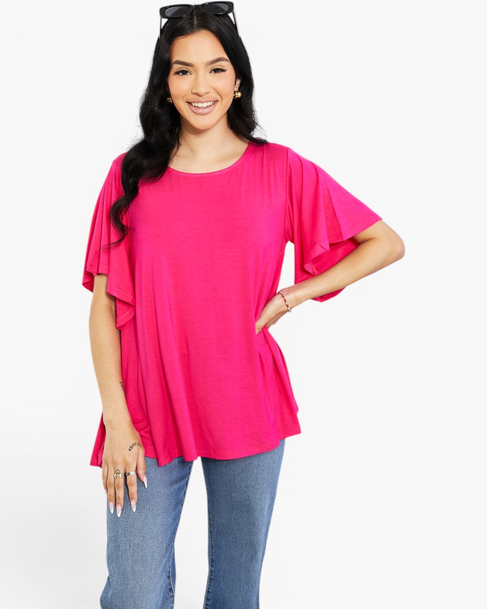 A woman modeling a bright pink short-sleeve top with layered ruffle details on the sleeves, paired with blue jeans, against a plain white studio background.