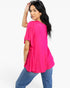 A woman modeling a bright pink short-sleeve top with a relaxed, draped silhouette and blue jeans, photographed against a clean white studio background.