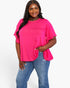 A woman modeling a bright pink short-sleeve top with a gathered, ruffled hem, paired with blue jeans, against a plain white studio background.