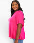 A woman wearing a bright pink short-sleeve top with a draped, fluttery hem detail, paired with blue jeans, photographed against a clean white background.