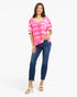 A woman modeling a pink and white tie-dye short-sleeve V-neck top with dark blue cropped jeans and white heeled sandals, photographed against a light gray studio background.