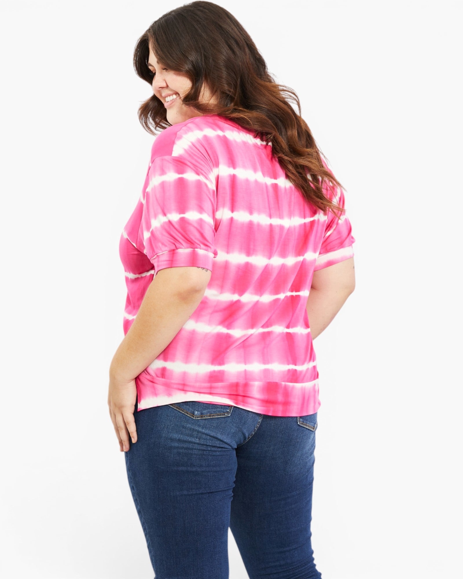 A woman wearing a pink and white tie-dye short-sleeve top and dark blue jeans, shown from a back-three-quarter angle against a plain white background.