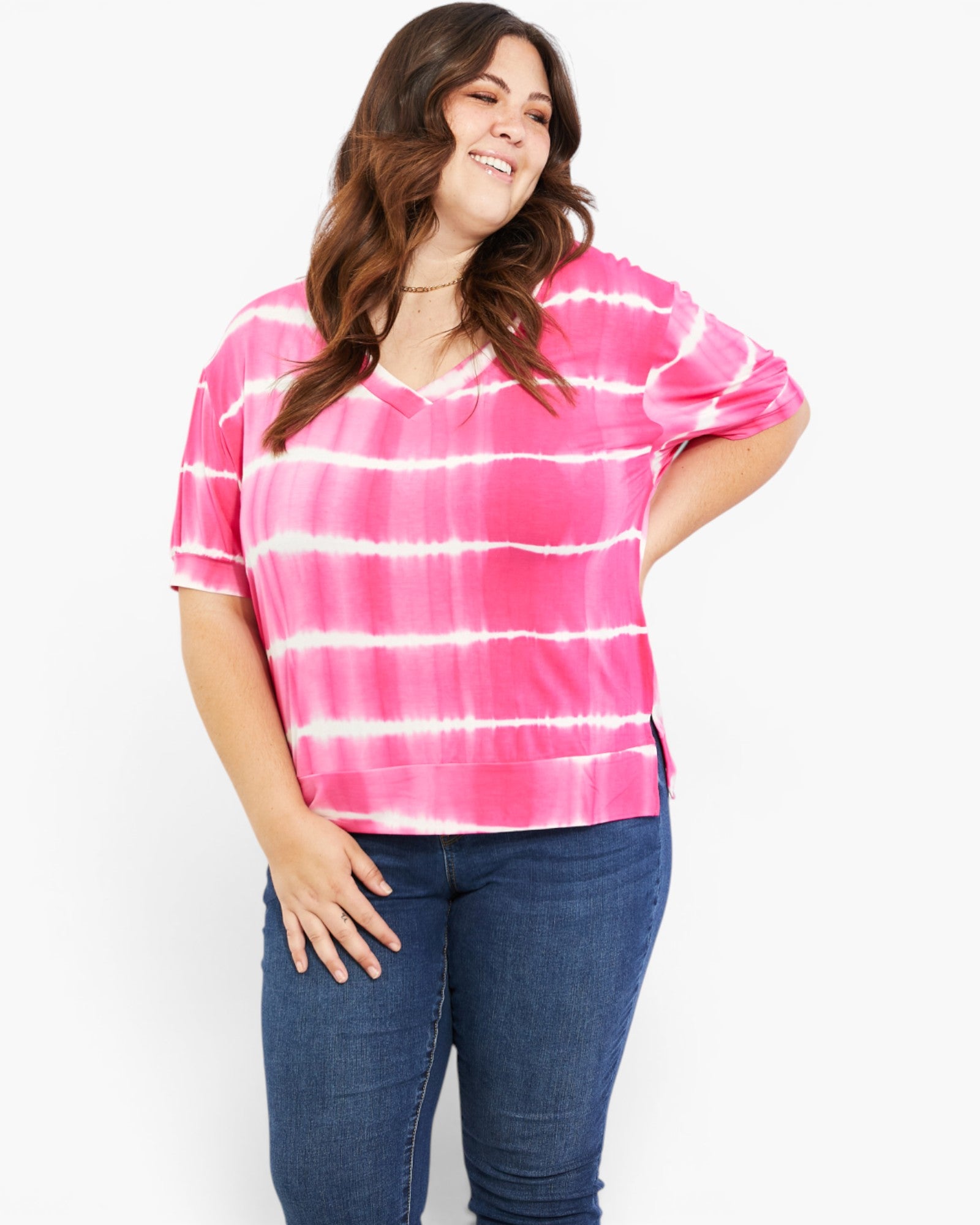 A woman modeling a pink and white tie-dye short-sleeve V-neck top with horizontal tie-dye bands, paired with dark blue jeans, against a plain white background.