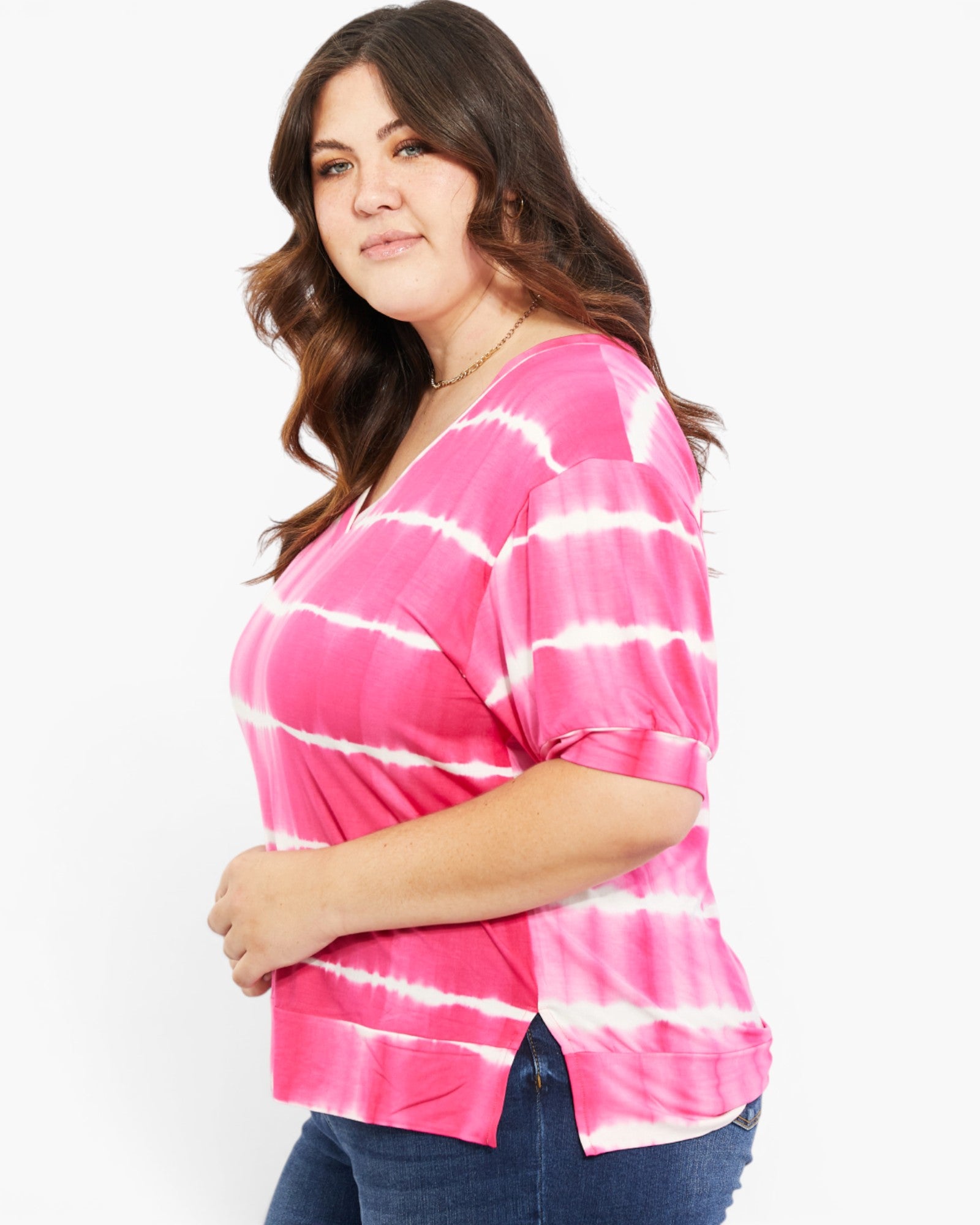A woman wearing a pink and white tie-dye short-sleeve top with rolled cuffs, paired with dark blue jeans, photographed against a plain white studio background.