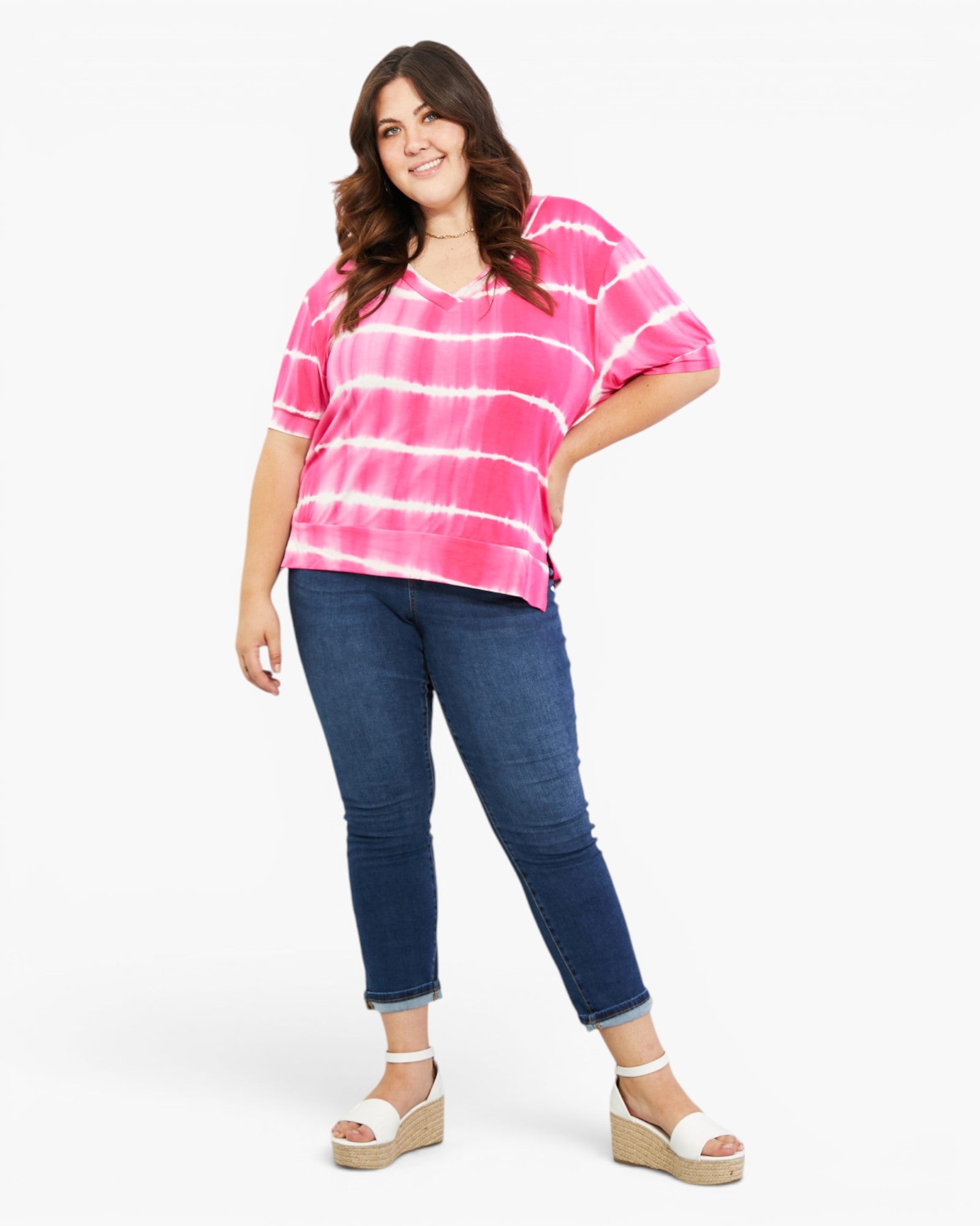 A woman modeling a bright pink and white tie-dye short-sleeve top with dark blue jeans and white wedge sandals, photographed against a clean white studio background.