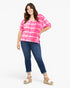 A woman modeling a bright pink and white tie-dye short-sleeve top with dark blue jeans and white wedge sandals, photographed against a clean white studio background.