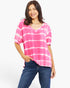 A woman wearing a pink and white tie-dye short-sleeve V-neck top with blue jeans, standing against a plain light gray studio background.