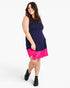 A woman modeling a sleeveless mini dress with a navy blue upper section and a bright pink ruffled hem, paired with black ankle boots, against a light gray studio background.