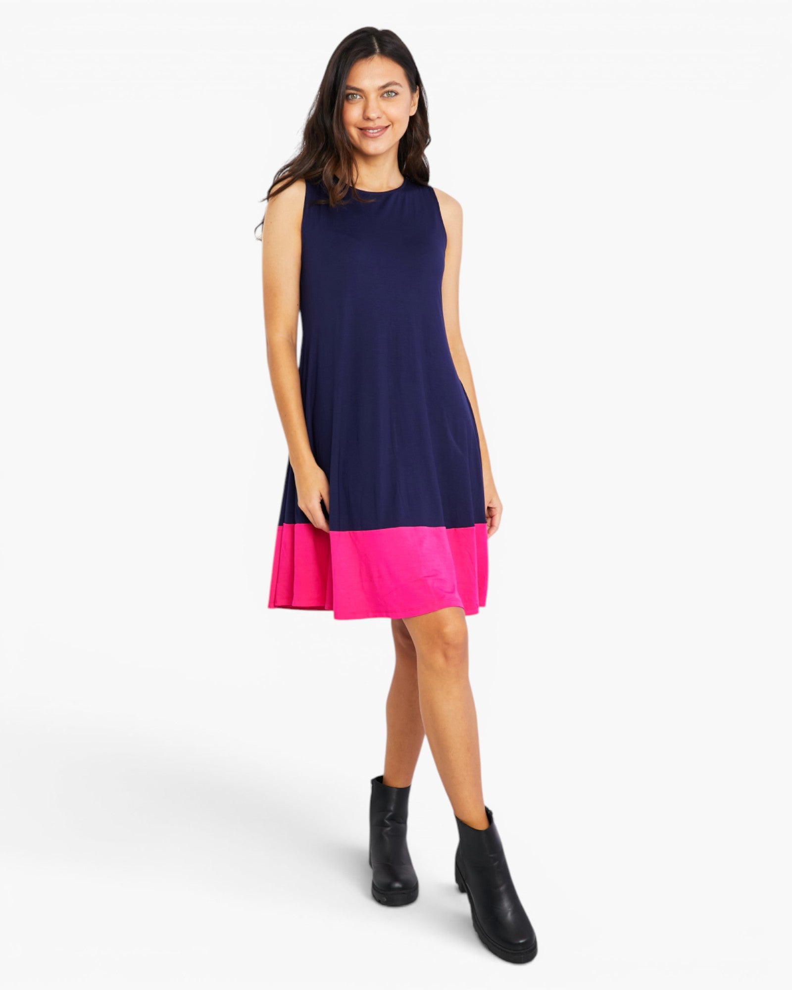 A woman modeling a sleeveless color-block mini dress with a navy blue upper section and a bright pink ruffled hem, paired with black ankle boots, against a light gray studio background.