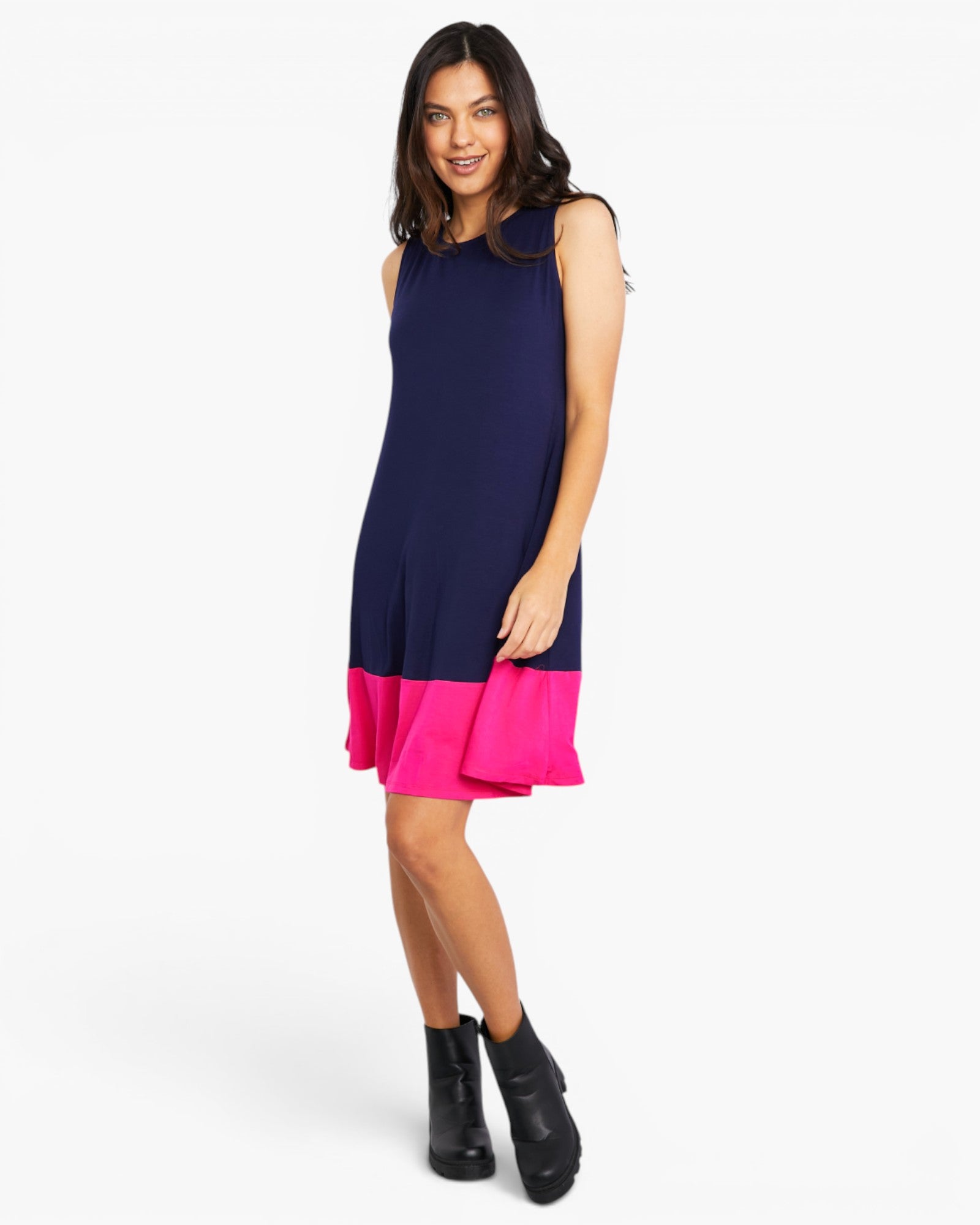 A woman modeling a sleeveless colorblock mini dress with a navy blue upper body and a bright pink ruffled hem, paired with black ankle boots, against a light gray studio background.