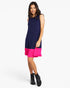 A woman modeling a sleeveless colorblock mini dress with a navy blue upper body and a bright pink ruffled hem, paired with black ankle boots, against a light gray studio background.