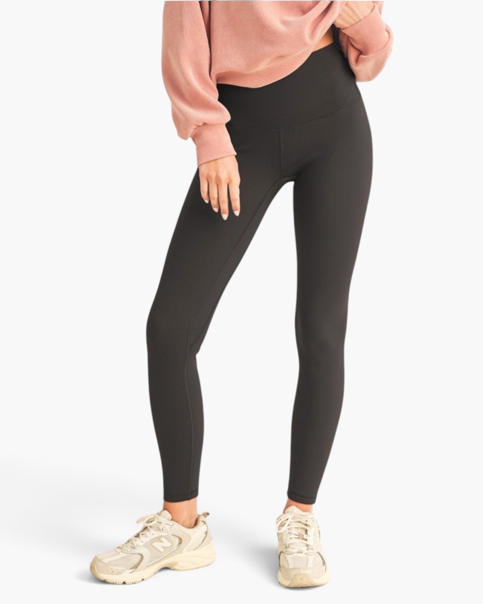 A model wearing high-waisted black leggings, a dusty pink sweatshirt, and beige sneakers, shown from the waist down against a light gray studio background.