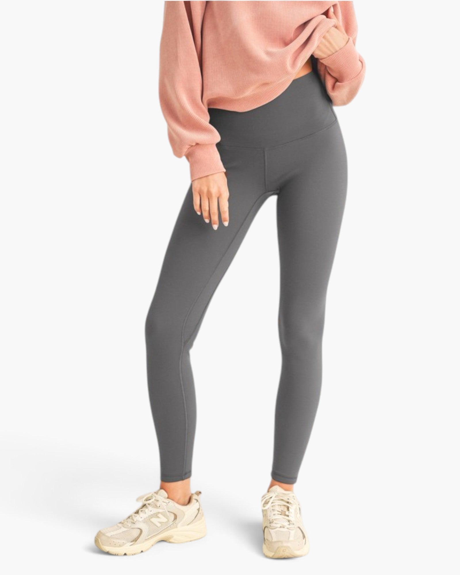 A model wearing high-waisted dark gray leggings, a dusty pink sweatshirt, and beige sneakers, shown from the waist down against a clean white background.