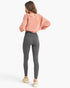 A woman shown from the back wearing high-waisted dark gray leggings, a dusty pink cropped sweatshirt, and beige sneakers against a plain white studio background.