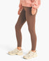 A model wearing brown leggings, a dusty pink sweatshirt, and beige New Balance sneakers, shown from the waist down against a light gray studio background.