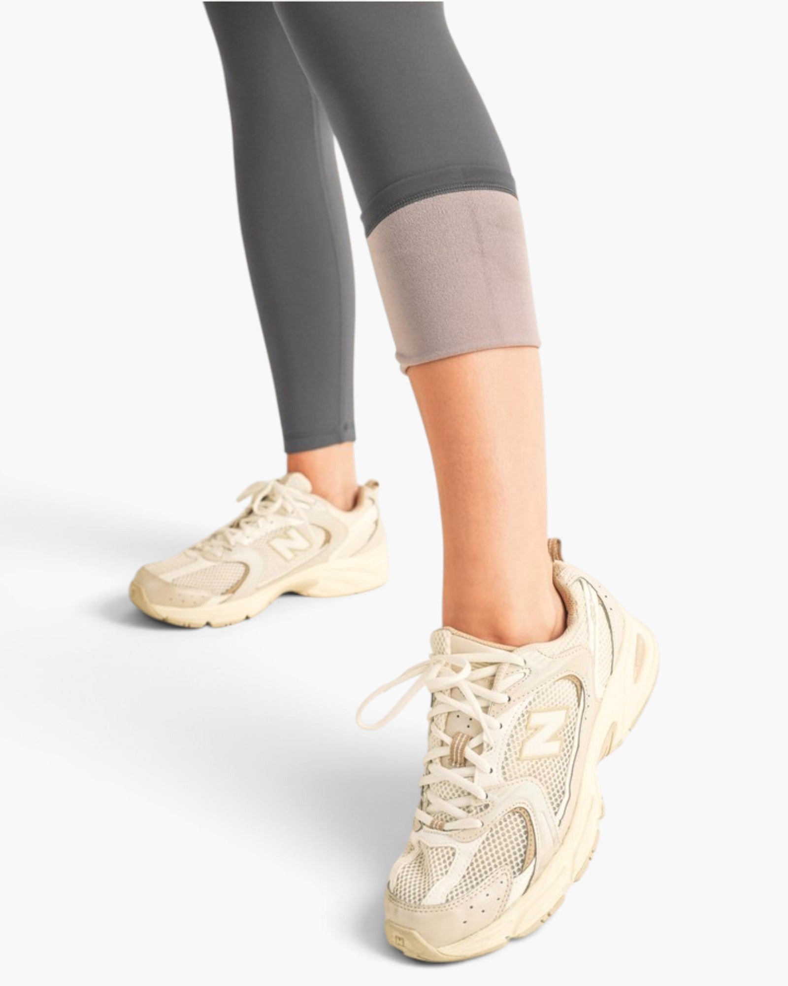 A person wearing beige New Balance chunky sneakers and dark gray leggings, shown from the knees down against a clean white studio background.