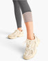 A person wearing beige New Balance chunky sneakers and dark gray leggings, shown from the knees down against a clean white studio background.