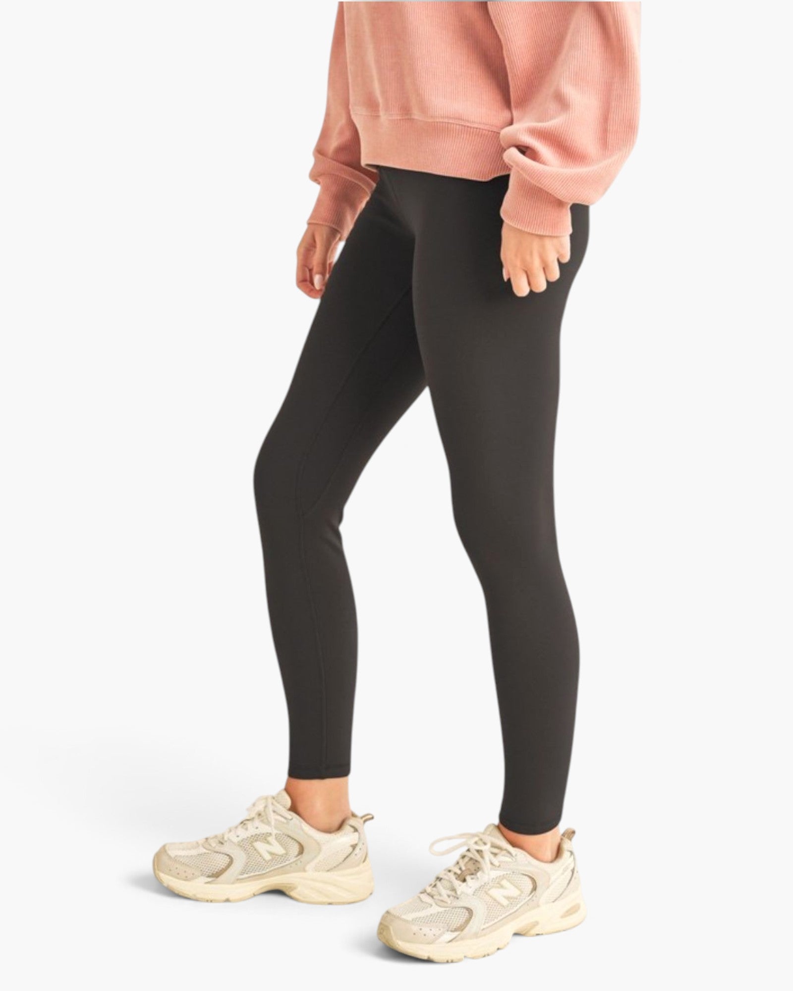 A model wearing black leggings, a dusty pink sweatshirt, and beige New Balance sneakers, shown from the waist down against a light gray studio background.