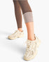 A person wearing beige New Balance sneakers and brown leggings with a gray compression cuff, shown from the knees down against a clean white studio background.