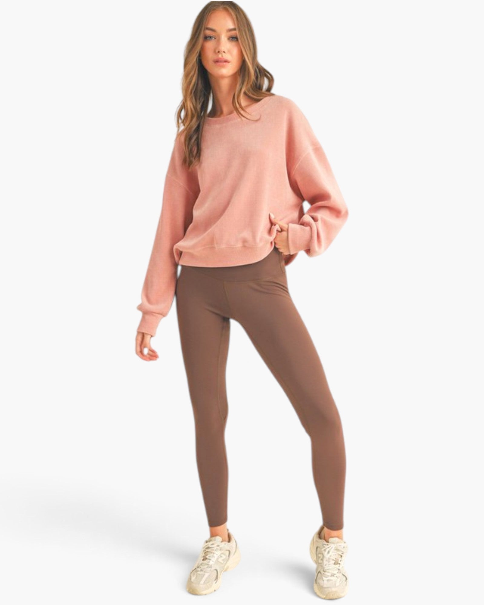 A woman modeling a dusty pink crewneck sweatshirt and brown leggings with white sneakers, standing against a light gray studio background.