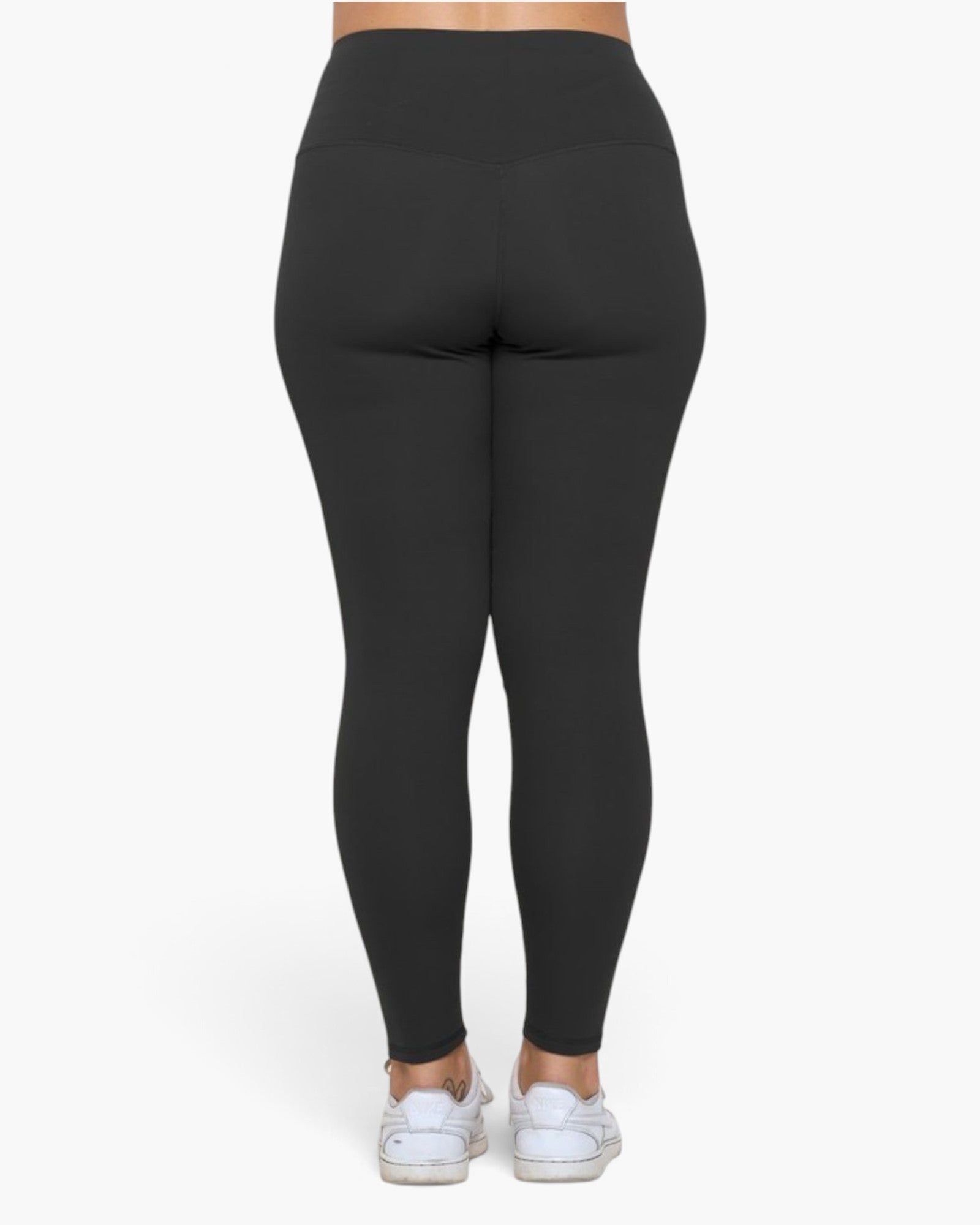 Rear view of a person wearing black high-waisted leggings and white sneakers against a plain light gray studio background.