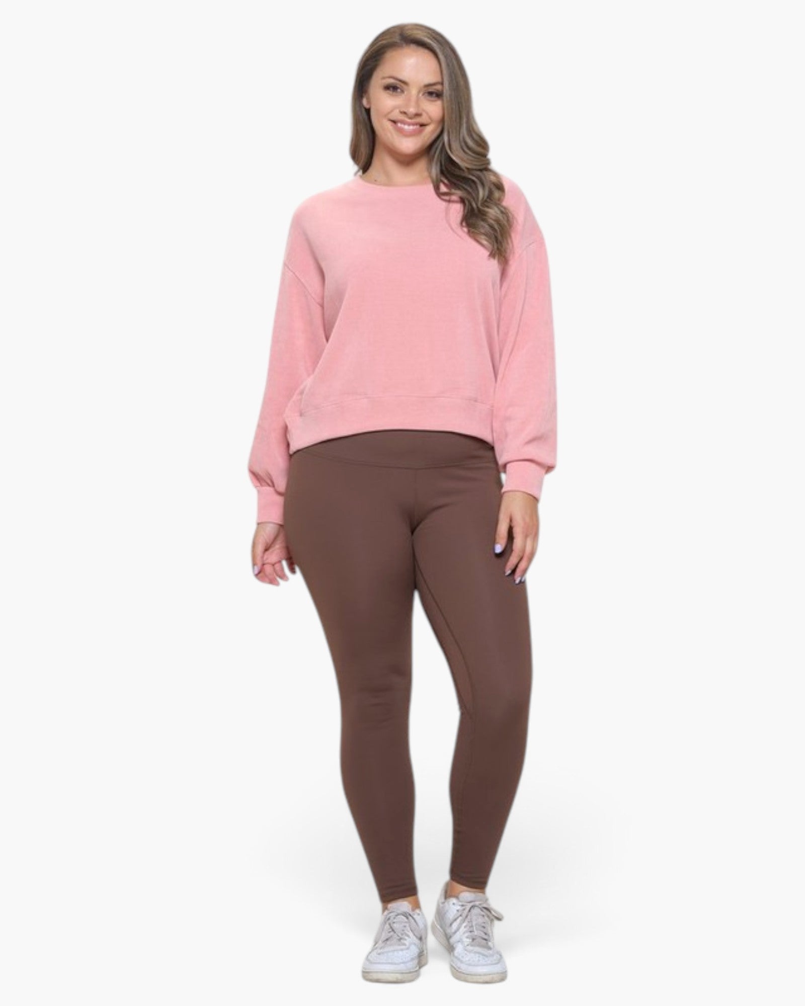 A woman modeling a light pink long-sleeve crewneck sweatshirt and brown leggings with white sneakers, standing against a plain white studio background.