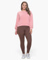 A woman modeling a light pink long-sleeve crewneck sweatshirt and brown leggings with white sneakers, standing against a plain white studio background.