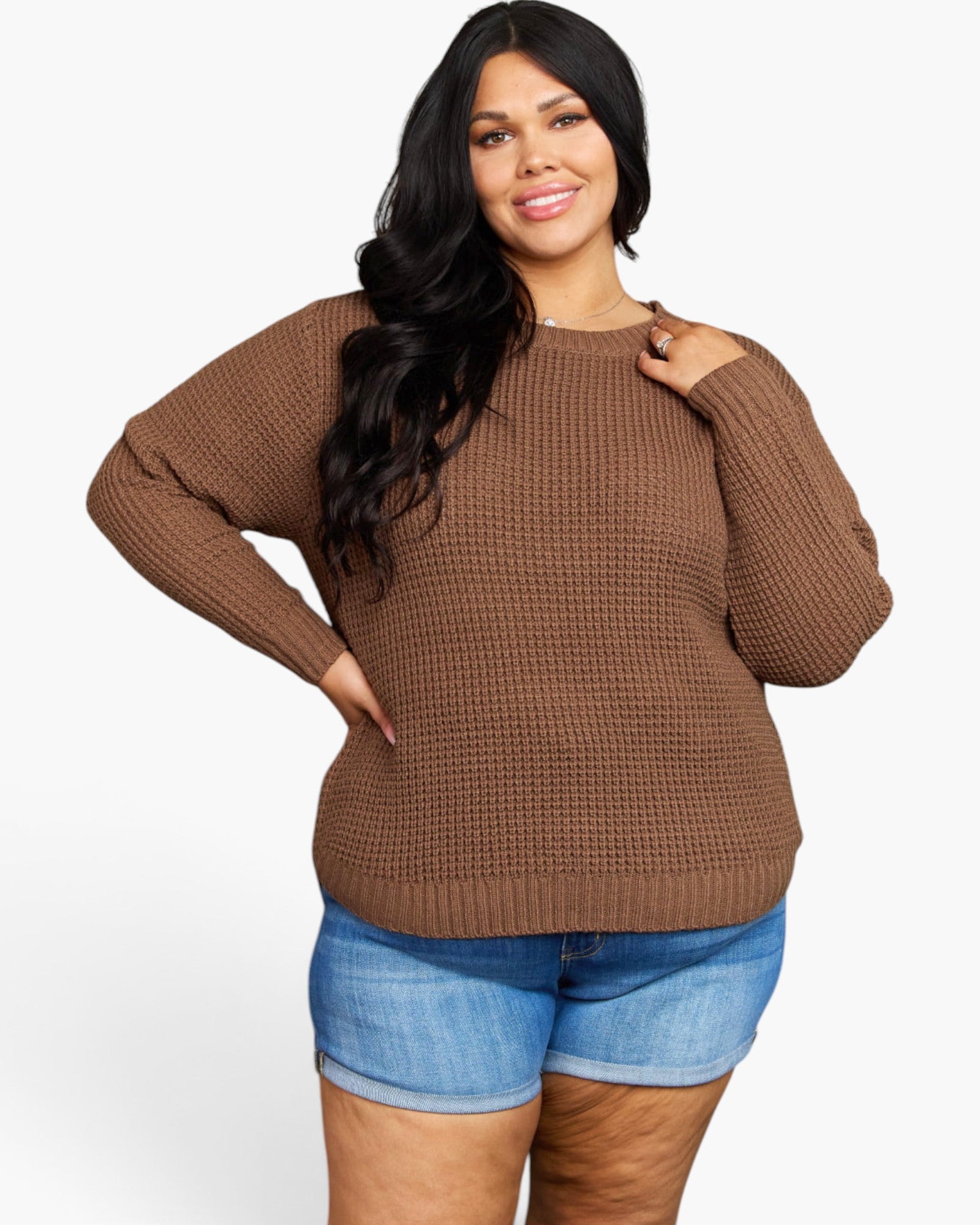 A woman modeling a brown waffle-knit long-sleeve sweater with a crew neck, paired with blue denim shorts, against a plain white background.