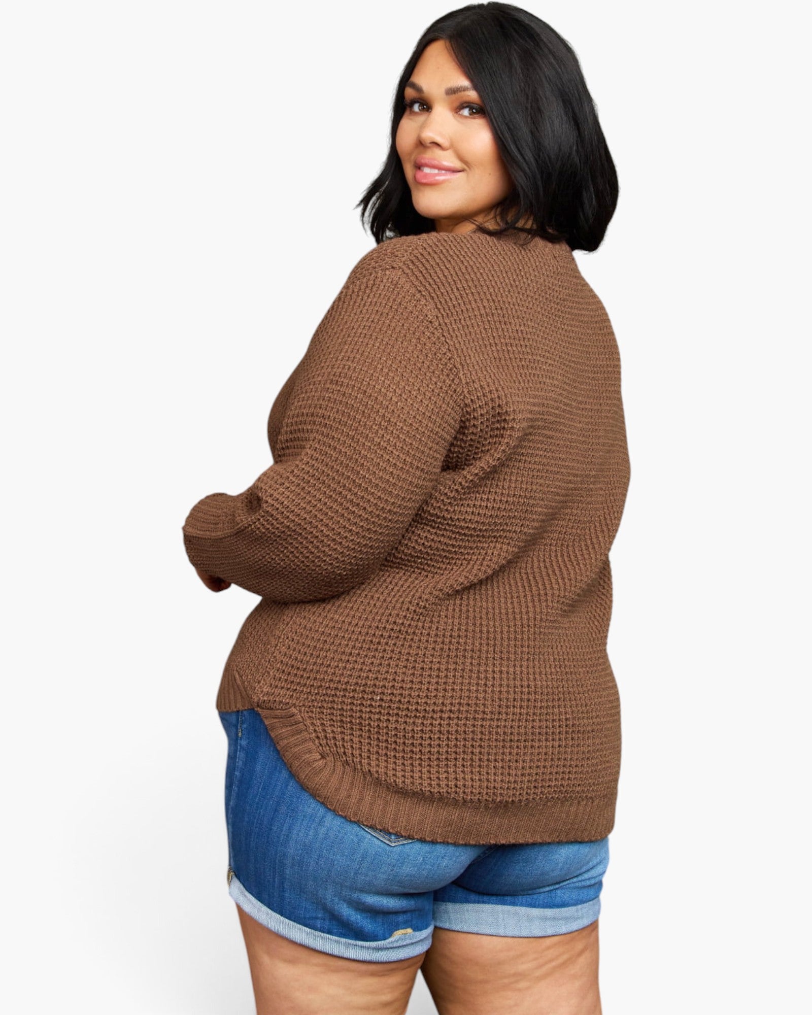 A woman wearing a brown waffle-knit sweater and blue denim shorts, photographed from the back against a plain light gray studio background.
