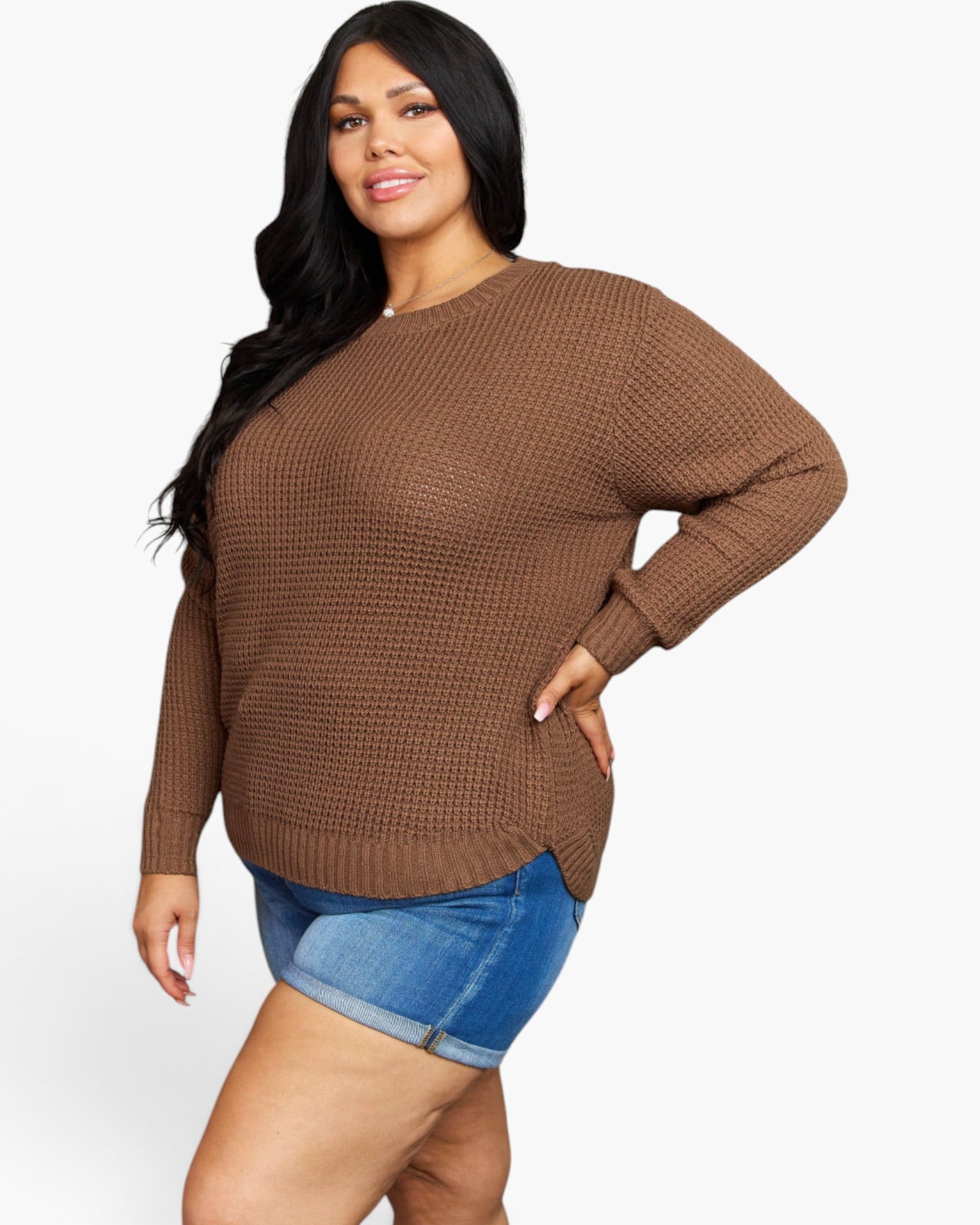 A woman modeling a brown waffle-knit long-sleeve sweater with a relaxed fit, paired with blue denim shorts, against a plain white studio background.