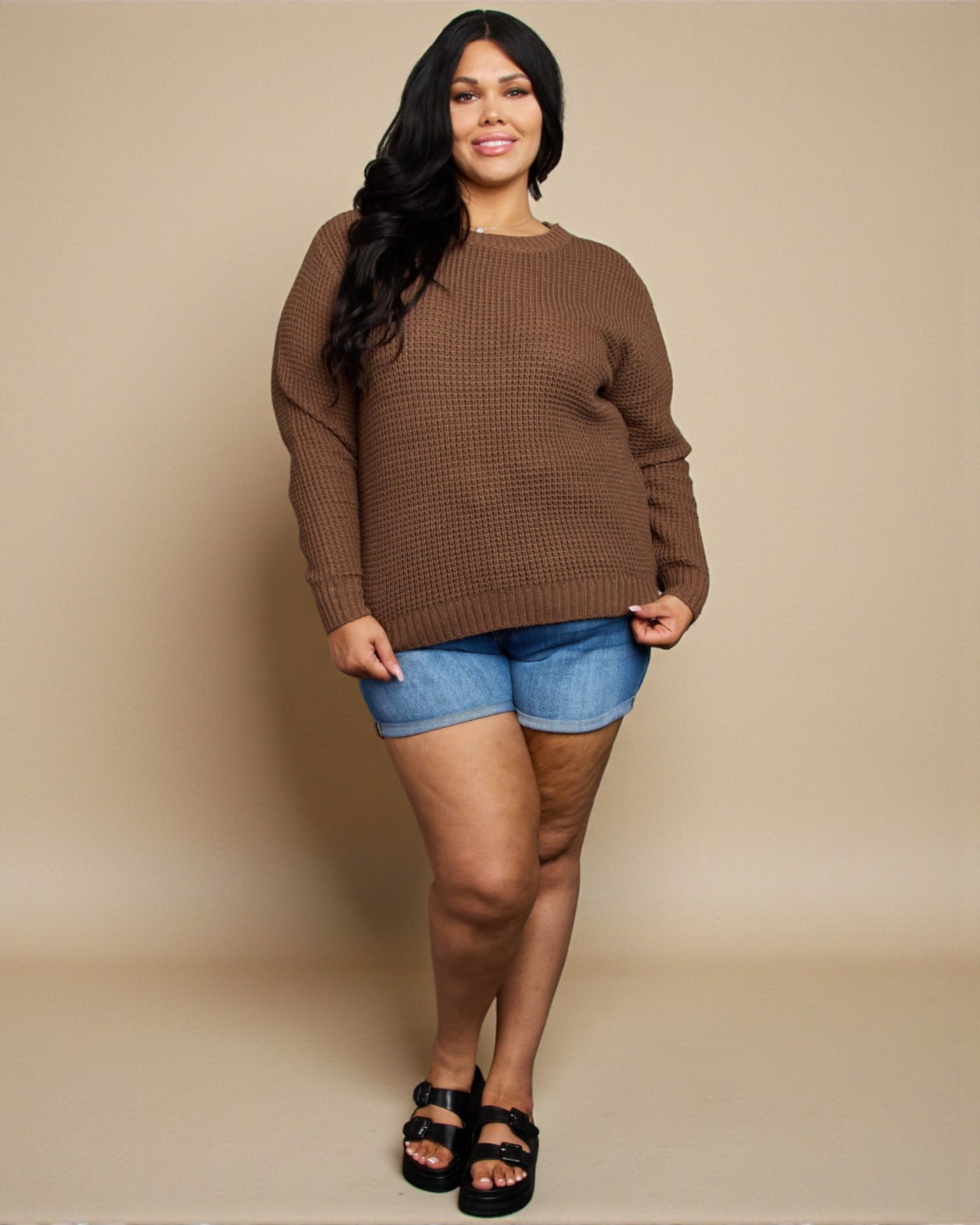 Woman wearing a brown sweater and denim shorts against a beige background