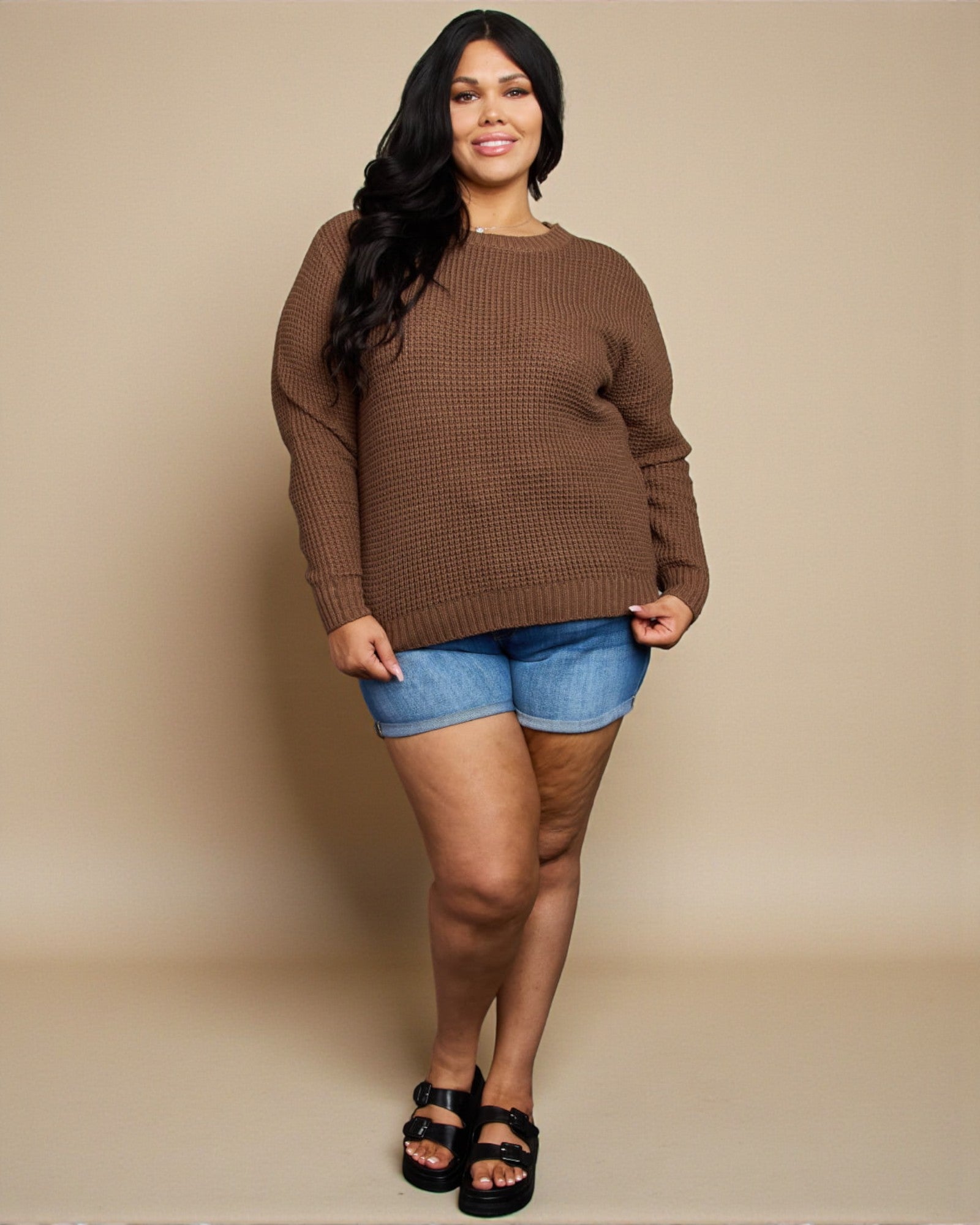 Woman wearing a brown sweater and denim shorts against a beige background