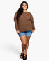 A woman modeling a brown waffle-knit sweater, paired with denim shorts and black sandals, against a plain white studio background.