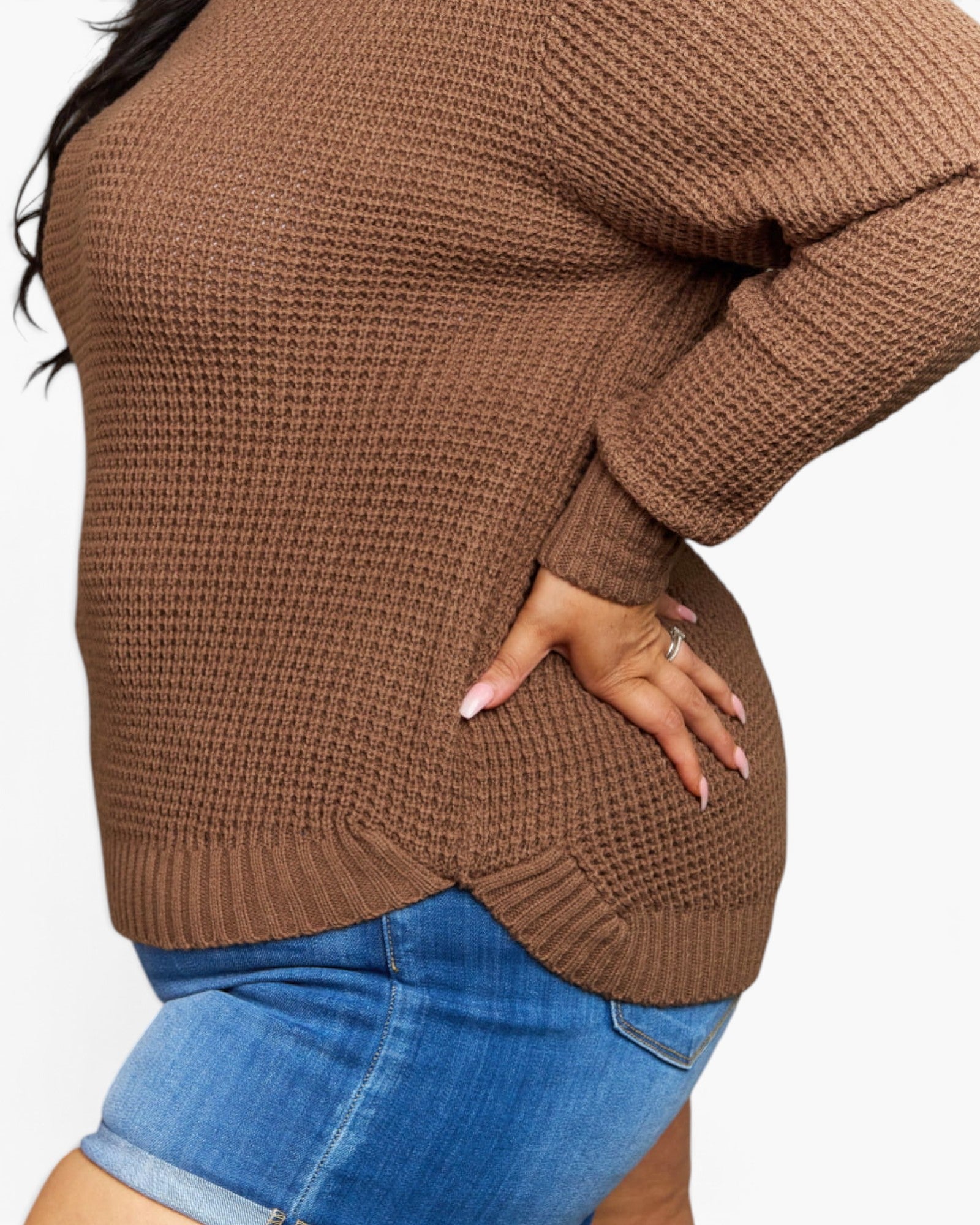 A close-up of a person wearing a brown ribbed knit sweater and blue denim shorts against a plain light background.