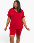 A woman modeling a matching red two-piece lounge set consisting of a short-sleeve V-neck top and fitted biker shorts, photographed against a plain light gray studio background.