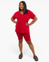 A woman modeling a matching red two-piece loungewear set consisting of a short-sleeve V-neck top and fitted biker shorts, paired with black slide sandals, against a plain white studio background.