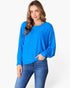 A woman wearing a bright blue long-sleeve knit top with a relaxed fit and blue jeans, standing against a light gray studio background.