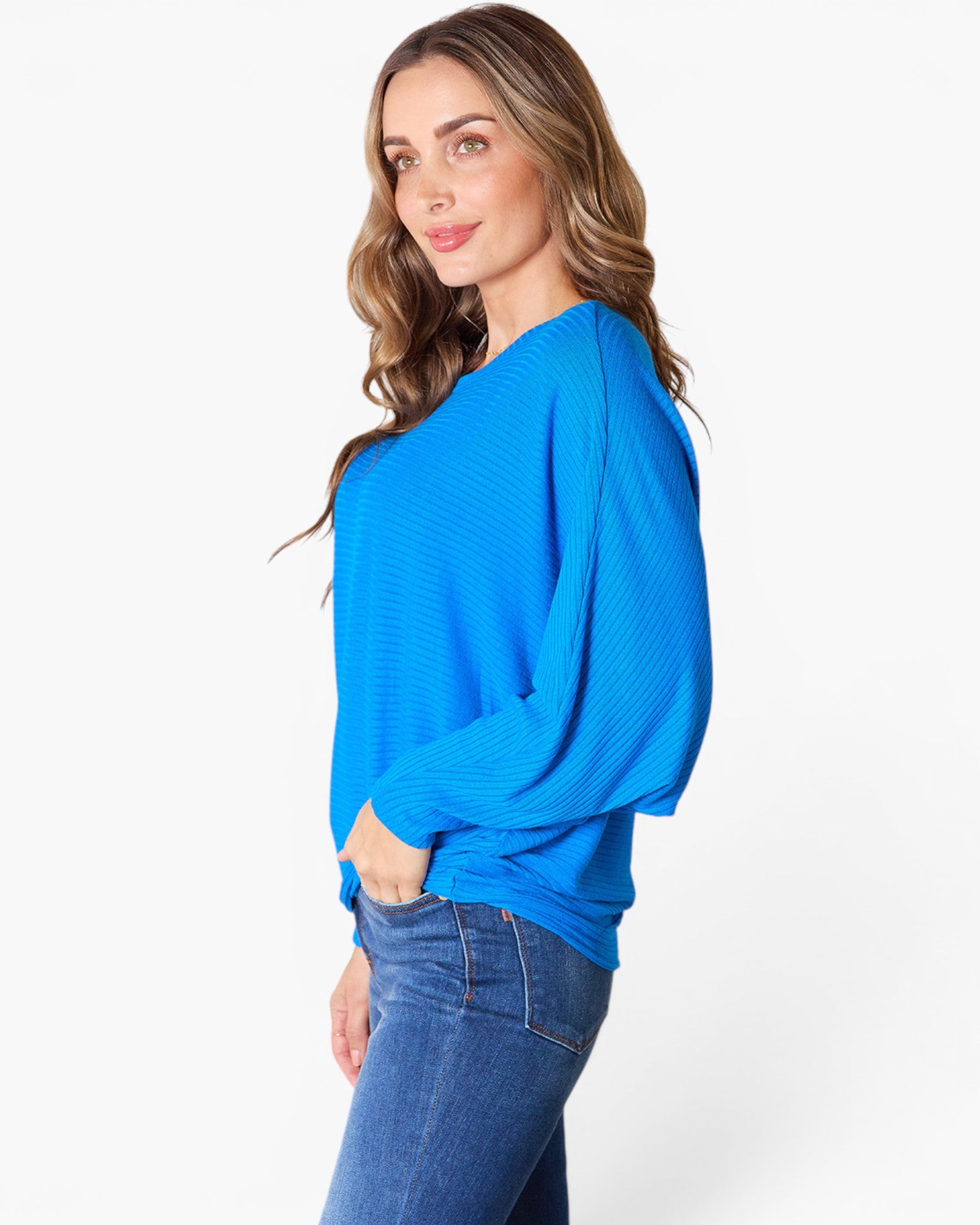 A woman wearing a bright blue long-sleeve knit top and blue jeans, photographed against a clean white studio background.