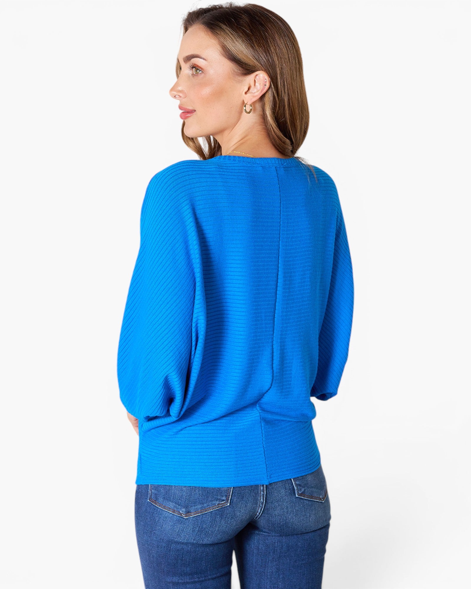 A woman shown from the back wearing a bright blue ribbed knit sweater and blue jeans against a plain light gray studio background.