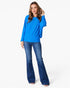 A woman modeling a bright blue long-sleeve knit top with flared legs, paired with dark blue jeans, against a plain white studio background.
