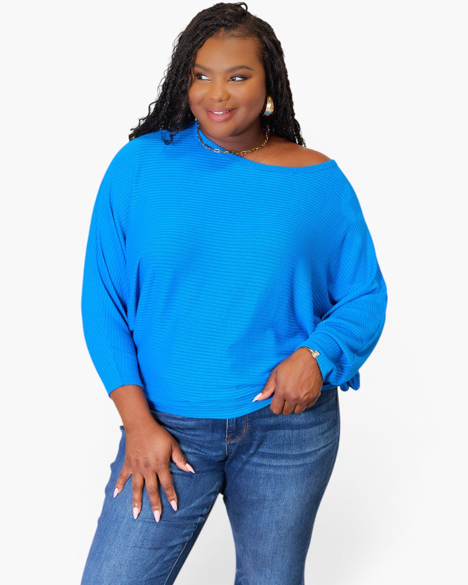 A woman modeling a bright blue off-the-shoulder knit top with long sleeves, paired with blue jeans, against a plain light gray background.
