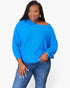 A woman modeling a bright blue off-the-shoulder knit top with long sleeves, paired with blue jeans, against a plain light gray background.