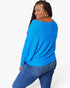 A woman modeling a bright blue long-sleeve knit top with a relaxed, draped fit and side knot detail, paired with blue jeans against a light gray studio background.